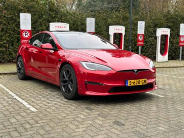 Een rode Tesla Model S Plaid staat geparkeerd bij een Tesla Supercharger-station met meerdere oplaadpunten omgeven door groen. Op het kenteken staat "S-628-GN.