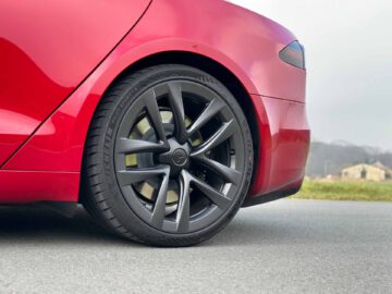 Close-up van de achterband en het wiel van een rode Tesla Model S Plaid geparkeerd op een verharde weg, met een onscherpe achtergrond van groen gras en huizen.