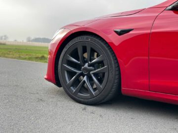 Close-up van het rode voorwiel en de band van een Tesla Model S Plaid op een verharde weg, met grasvelden en een bewolkte lucht op de achtergrond.