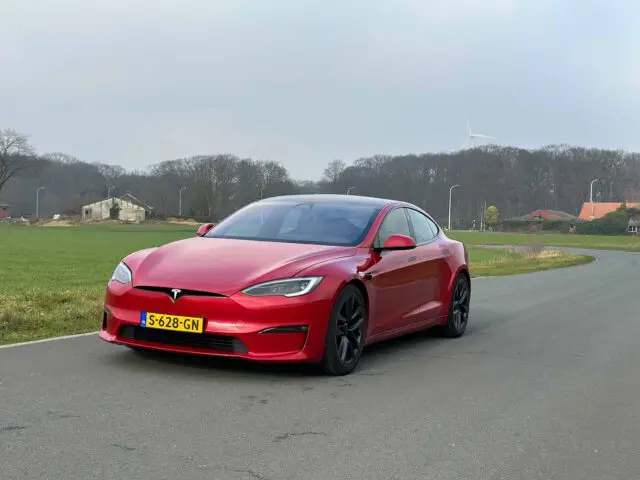 Een rode Tesla Model S Plaid staat geparkeerd op een gebogen landelijke weg, met een veld, bomen en een windturbine op de achtergrond.