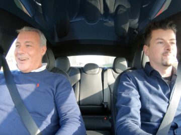 Op de voorstoelen van een Tesla Model S Plaid zitten twee mannen, beiden met veiligheidsgordels om. De man links lacht, terwijl de man rechts aandachtig vooruitkijkt. Het interieur van de auto is modern en goed verlicht.