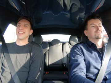 Twee mensen zitten in een Tesla Model S Plaid. De persoon links lacht, terwijl de bestuurder rechts gefocust is op de weg. Beiden dragen veiligheidsgordels en de achtergrond toont een heldere, zonnige dag.