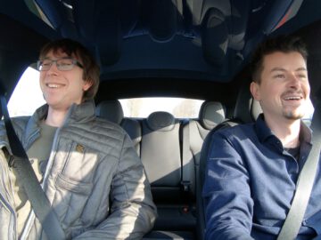 Twee mannen zitten glimlachend op de voorstoelen van een Tesla Model S Plaid. De ene man draagt een bril en een grijze jas, terwijl de ander een donkerblauwe jas draagt. Beiden dragen veiligheidsgordels.