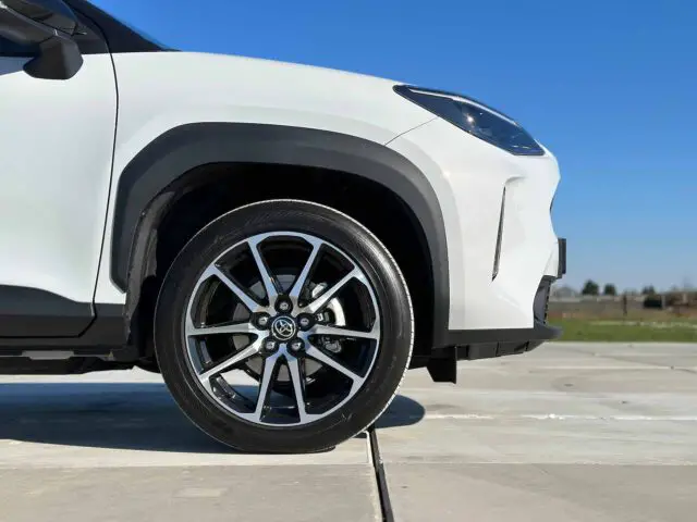 Een close-up van het linkervoorwiel van een witte Toyota Yaris Cross GR Sport, geparkeerd op een betonnen ondergrond onder een helderblauwe lucht.