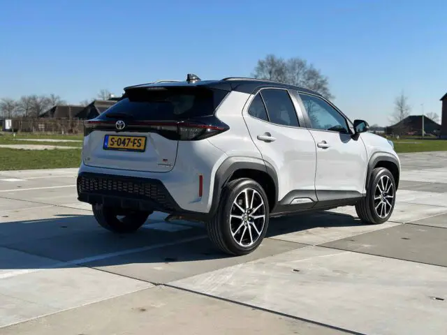 Een witte Toyota Yaris Cross GR Sport SUV staat geparkeerd op een verhard oppervlak met helder weer en gebouwen op de achtergrond.