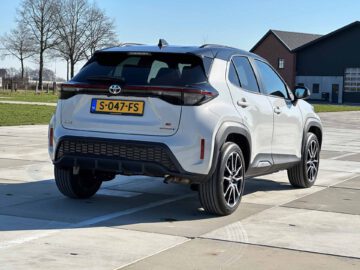 Een witte Toyota Yaris Cross GR Sport Hybrid staat geparkeerd op een verhard terrein met gebouwen en bomen op de achtergrond.