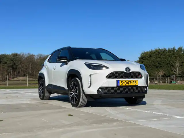 Een strakke witte Toyota Yaris Cross GR Sport SUV staat buiten geparkeerd op een verhard terrein met bomen en een helderblauwe lucht op de achtergrond. Op het kenteken aan de voorkant staat "S-047-FS.