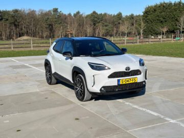 Een witte Toyota Yaris Cross GR Sport SUV staat buiten geparkeerd op een groot betonnen terrein, met op de achtergrond een bosgebied zichtbaar. Het voertuig heeft een Nederlands kenteken S-047-FS.