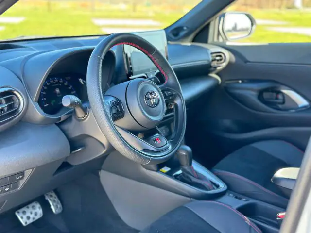 Interieur van de Toyota Yaris Cross GR Sport met een zwart en rood stuur, digitaal dashboarddisplay en touchscreen-infotainmentsysteem. Het strakke interieur wordt aangevuld met toegankelijke bedieningsknoppen.