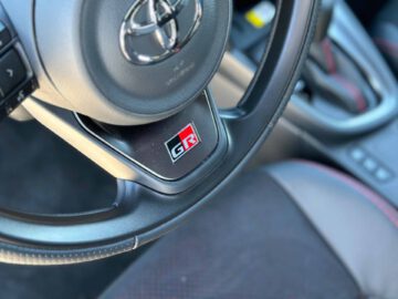 Close-up van een Toyota-autostuur met een 'GR'-embleem in het midden, dat de sportieve elegantie van de Toyota Yaris Cross GR Sport laat zien, met een deel van het interieur van de auto zichtbaar op de achtergrond.