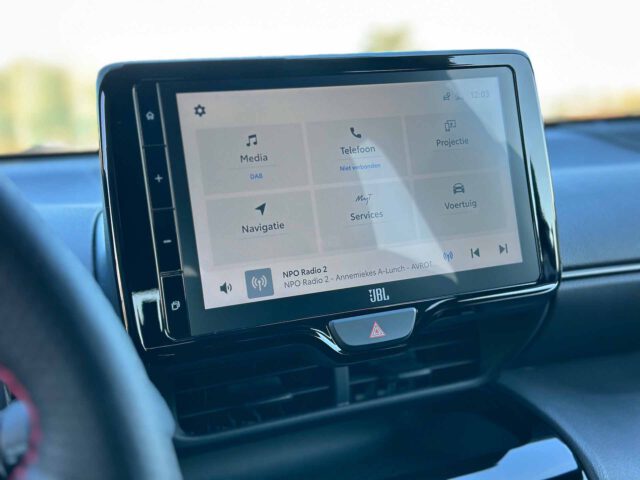 Close-up van het infotainmentsysteem van een Toyota Yaris Cross GR Sport, met een touchscreeninterface met opties voor media, telefoon, navigatie, services en voertuig. Onder het scherm bevindt zich een op het dashboard gemonteerde bedieningsknop.