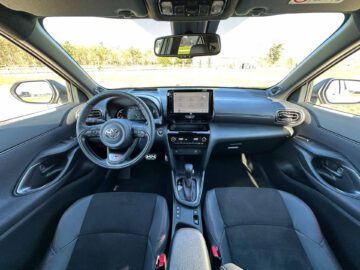 Binnenaanzicht van een moderne Toyota Yaris Cross GR Sport met een stuur met bedieningselementen, centraal touchscreen, versnellingspook en zwarte stoelen met rode stiksels.