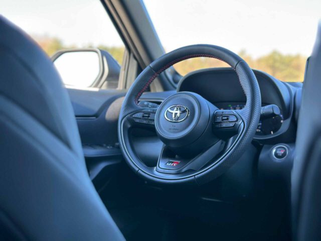 Close-up van het interieur van een Toyota Yaris Cross GR Sport, met het stuur, een deel van het dashboard en een zijspiegel op de achtergrond.