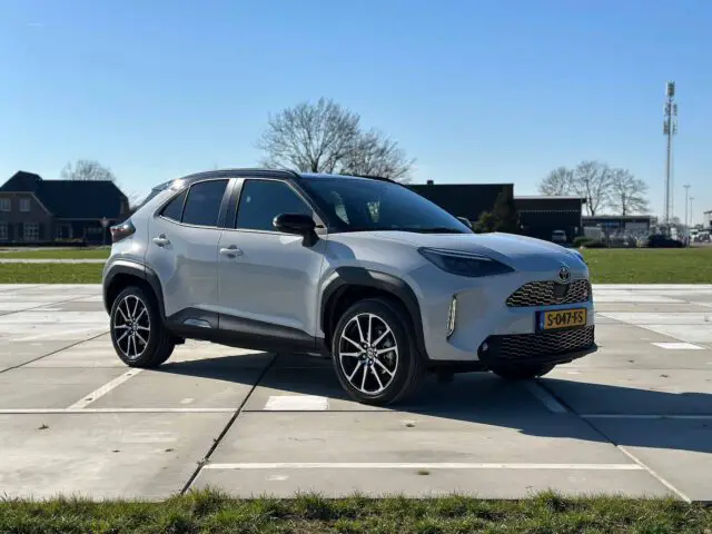 Een zilveren Toyota Yaris Cross GR Sport SUV staat geparkeerd op een verhard terrein met gras op de voorgrond en bomen en gebouwen op de achtergrond, onder een helderblauwe lucht.