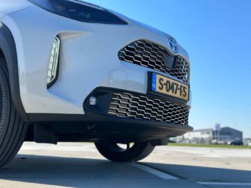 Close-up van de voorkant van een zilveren Toyota Yaris Cross GR Sport met een zichtbaar kenteken "S-047-FS." De auto staat geparkeerd op een betonnen ondergrond met een helderblauwe lucht en gebouwen op de achtergrond.