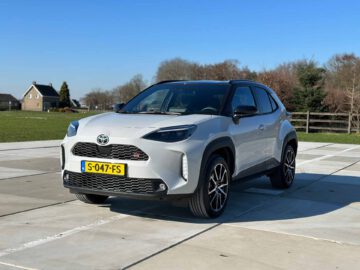 Een zilveren Toyota Yaris Cross GR Sport staat buiten geparkeerd op een betonnen ondergrond, met een helderblauwe lucht en bomen op de achtergrond. Aan de voorzijde is het Nederlandse kenteken S-047-FS zichtbaar.