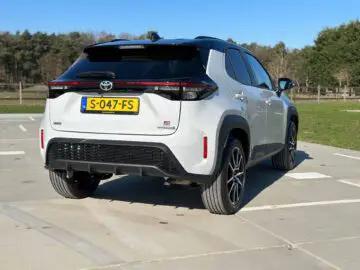 Een witte Toyota Yaris Cross GR Sport SUV met een hybride badge staat op een zonnige dag buiten geparkeerd. Het voertuig heeft een Europees kenteken "S-047-FS" en staat op een lege parkeerplaats met gras en bomen op de achtergrond.