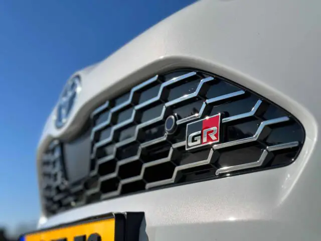 Close-up van een autogrille met een Toyota-embleem aan de linkerkant en een GR-badge aan de rechterkant, met de sportieve Toyota Yaris Cross GR Sport, met een deel van een kentekenplaat zichtbaar aan de onderkant.