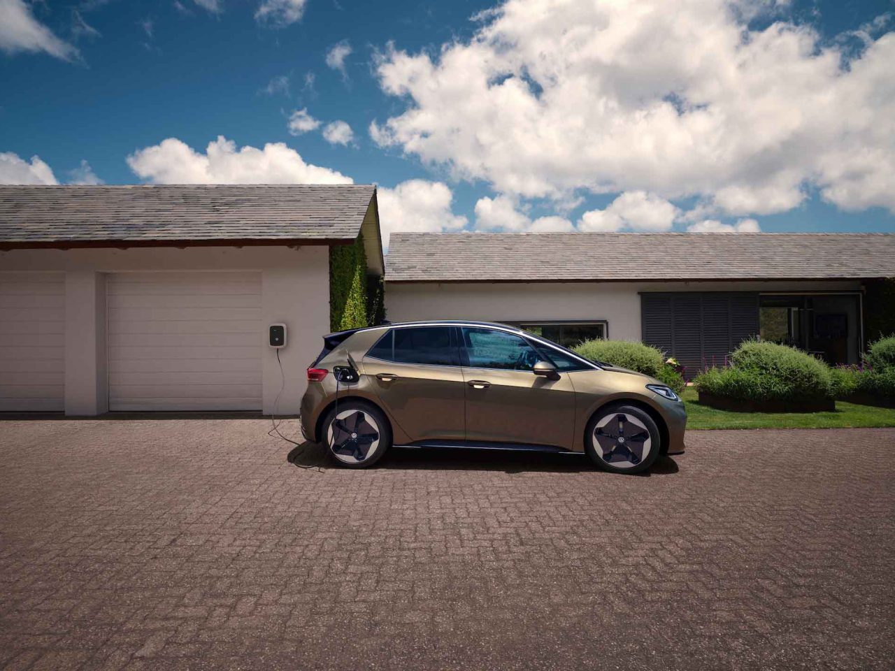 Een moderne gouden Volkswagen ID.3 elektrische auto staat geparkeerd op een oprit, aangesloten op een laadstation dat is bevestigd aan een witte garage van een huis met één verdieping onder een blauwe lucht met wolken.