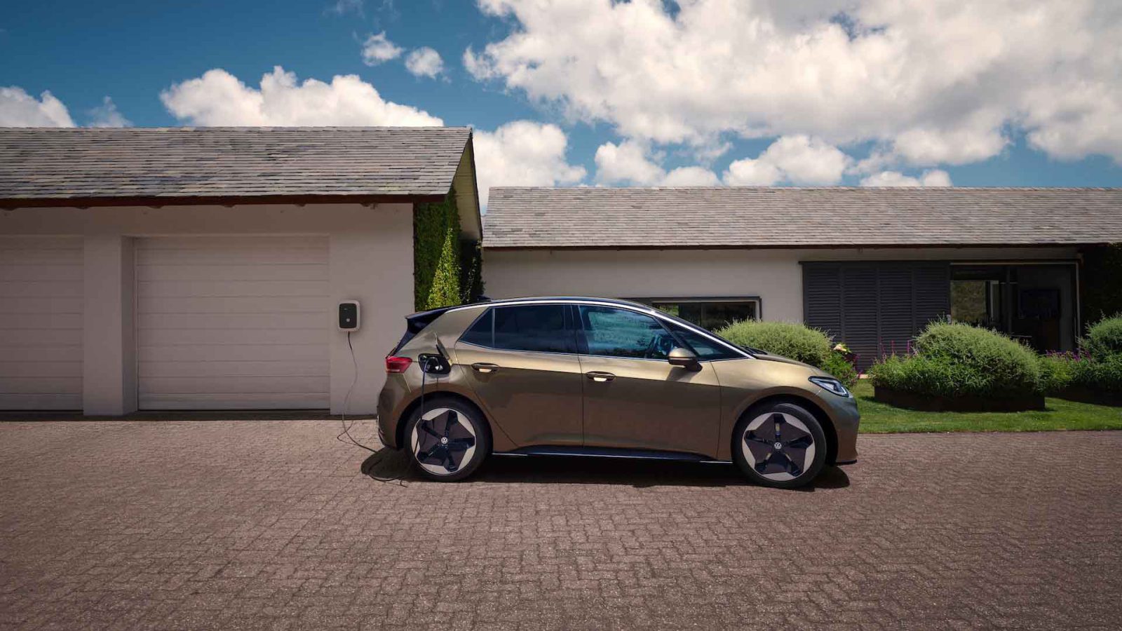 Een moderne gouden Volkswagen ID.3 elektrische auto staat geparkeerd op een oprit, aangesloten op een laadstation dat is bevestigd aan een witte garage van een huis met één verdieping onder een blauwe lucht met wolken.