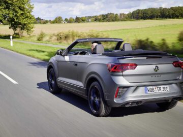 Een grijze Volkswagen T-Roc Cabrio rijdt over een landelijke weg, met de kap naar beneden, omgeven door velden en bomen onder een heldere hemel.