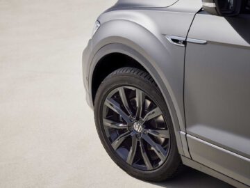 Close-up van de linkervoorhoek van een zilveren Volkswagen T-Roc Cabrio, gericht op het stuur en de band tegen een betonnen achtergrond.