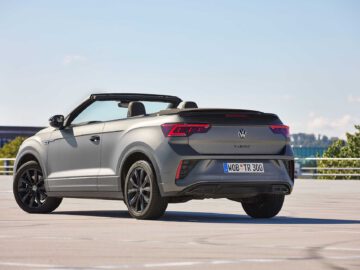 Een grijze Volkswagen T-Roc Cabrio staat buiten geparkeerd met een heldere lucht op de achtergrond. De auto is voorzien van zwarte velgen en een zichtbaar kenteken aan de achterzijde.