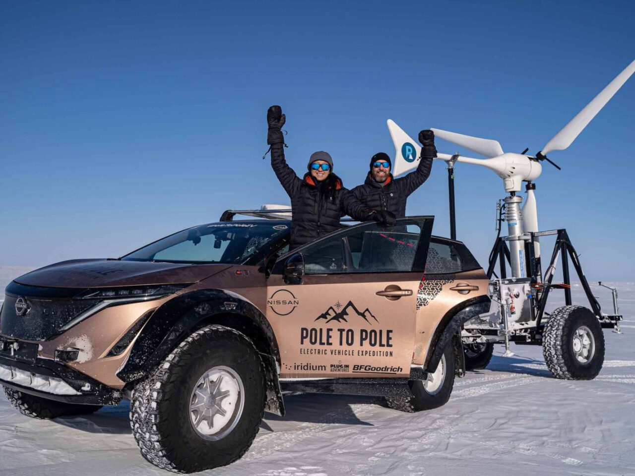 Twee mensen in winterkleding staan naast een elektrische auto van Nissan en een windturbinetrailer in een besneeuwd landschap feest te vieren. De auto draagt het merk "Pole to Pole Electric Vehicle Expedition".