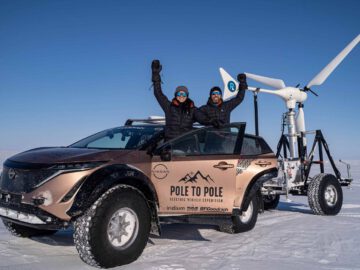 Twee mensen in winterkleding staan naast een elektrische auto van Nissan en een windturbinetrailer in een besneeuwd landschap feest te vieren. De auto draagt het merk "Pole to Pole Electric Vehicle Expedition".