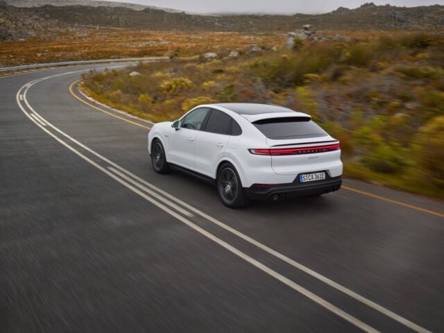 Een strak witte Porsche sportsedan rijdt over een bochtige weg in een heuvelachtig landschap met schaarse begroeiing.