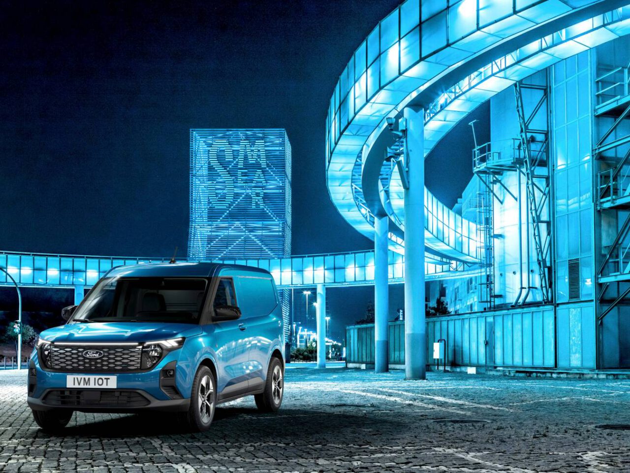 Een blauwe Ford E-Transit Courier staat geparkeerd op een verhard oppervlak in een moderne, verlichte stedelijke omgeving met futuristische, blauw verlichte constructies.
