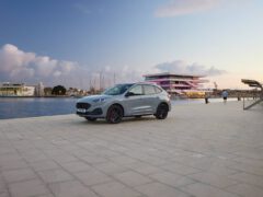 Een zilveren Ford Kuga staat geparkeerd aan de rand van een verhard gebied aan het water met een modern gebouw en aangemeerde boten op de achtergrond bij zonsondergang. Er lopen twee mensen op het nabijgelegen pad.