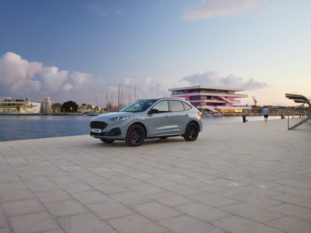 Een zilveren Ford Kuga staat geparkeerd aan de rand van een verhard gebied aan het water met een modern gebouw en aangemeerde boten op de achtergrond bij zonsondergang. Er lopen twee mensen op het nabijgelegen pad.