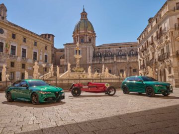 Drie auto's, waaronder twee moderne groene auto's en een vintage rode Alfa Romeo, worden tentoongesteld op een historisch Europees plein met een fontein en een sierlijk gebouw onder een helderblauwe lucht.