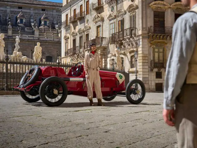 Een persoon in vintage racekleding staat bij een rode Alfa Romeo op een historisch plein met beelden en oude gebouwen op de achtergrond. Een andere persoon kijkt vanaf de rechterkant toe.