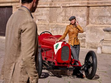 Een vrouw met een bruin jasje en een grijze hoed staat naast een rode vintage Alfa Romeo-raceauto, terwijl een man in een beige jasje naar haar kijkt in een buitenomgeving met rustieke gebouwen op de achtergrond.