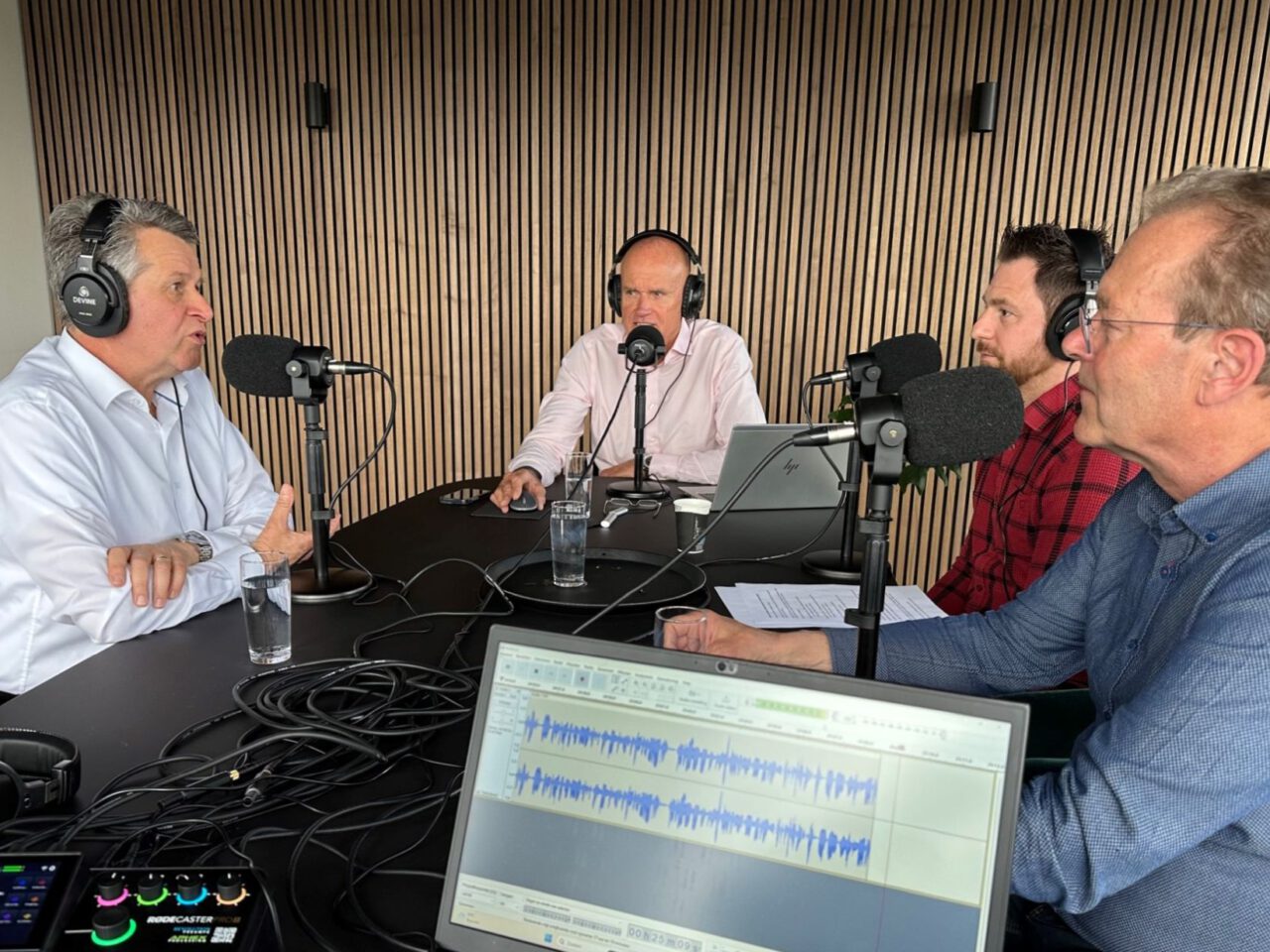 Vier mannen zitten aan een tafel met microfoons en koptelefoons en nemen een podcast of interview op. Op de tafel staan audioapparatuur en een laptop zichtbaar, waar mogelijk onderwerpen worden besproken die een BPM-verhoging bij hun luisteraars kunnen veroorzaken.