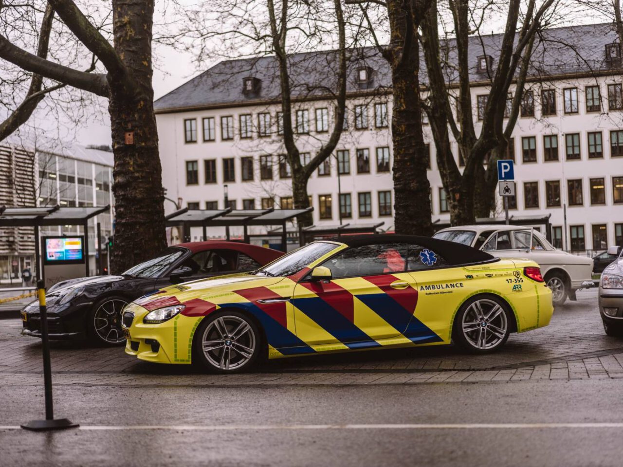 Een geel en rood gestreepte BMW-ambulance staat op een bewolkte dag naast andere voertuigen geparkeerd in een straat, met gebouwen en bomen op de achtergrond.