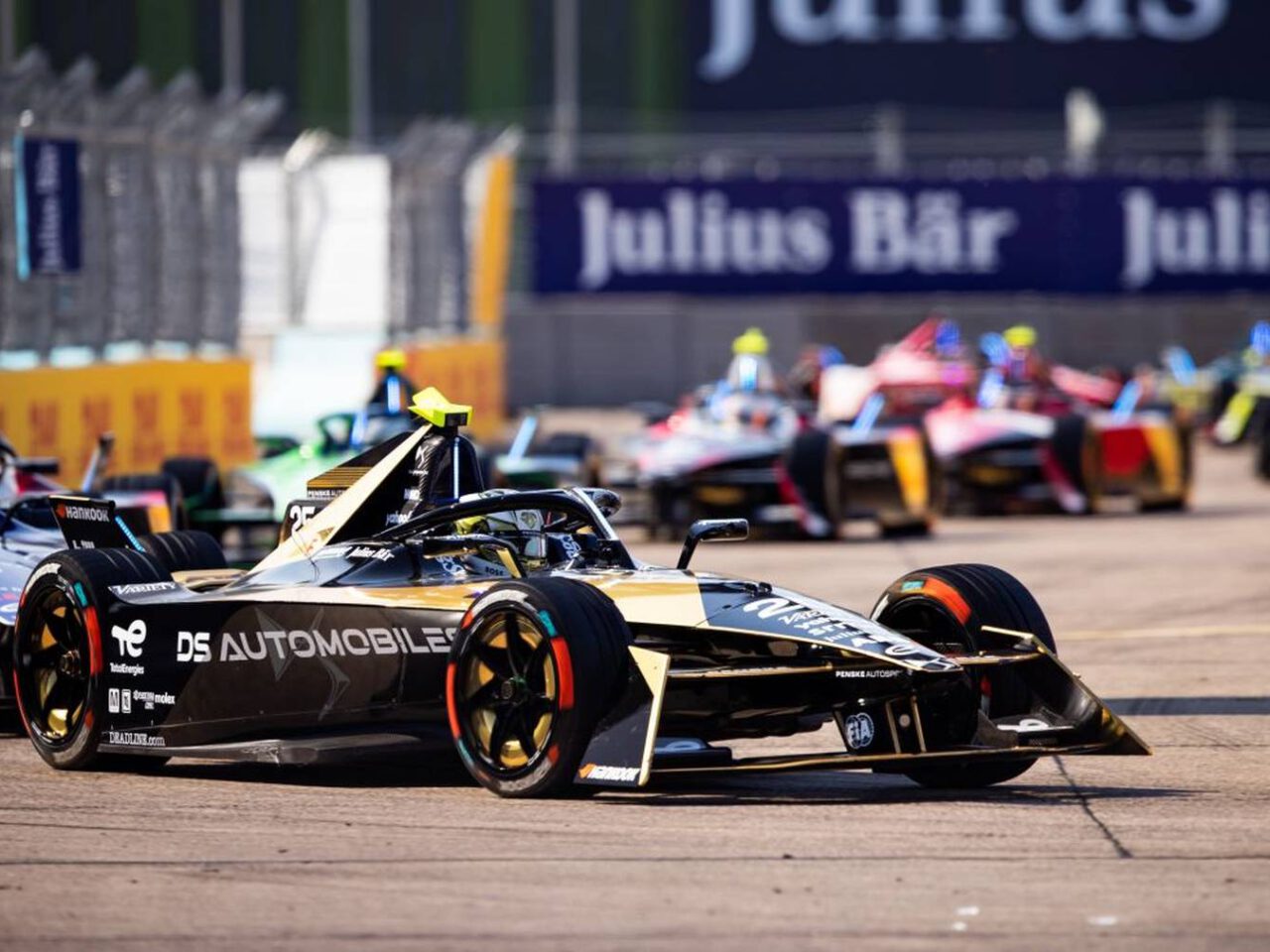 Elektrische raceauto's zoeven over het circuit tijdens een Formule E-race, met een auto van DS Automobiles voorop. Op de achtergrond zijn reclamebanners zichtbaar.