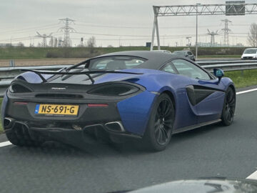 Een blauw-zwarte McLaren-sportwagen met achtervleugel rijdt op een snelweg en heeft kenteken NS-691-G. Op de achtergrond zijn bovengrondse borden en elektriciteitsleidingen zichtbaar.