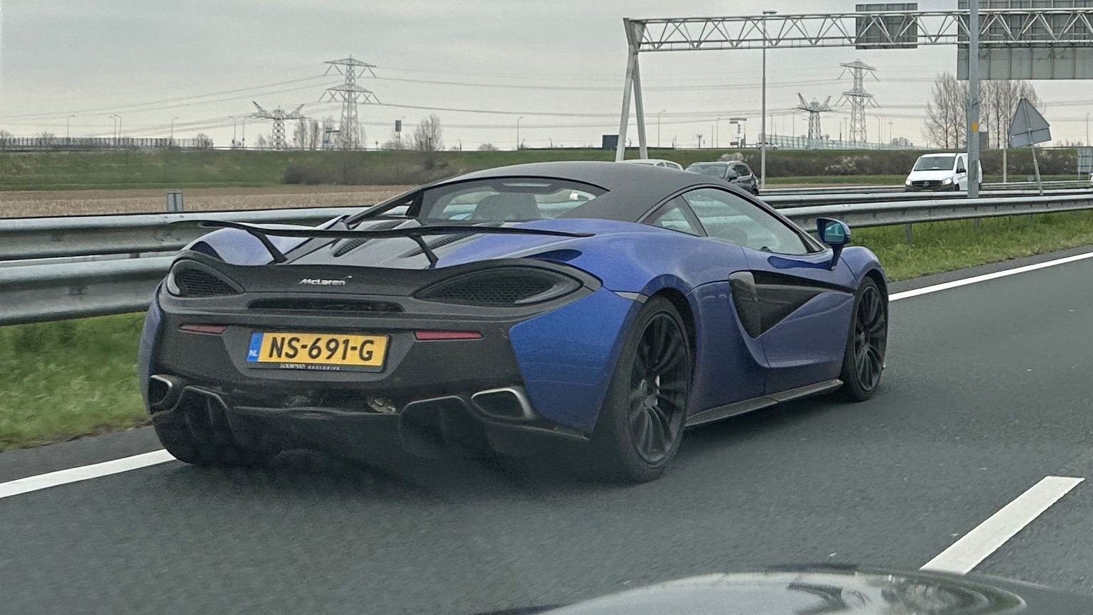 Een blauw-zwarte McLaren-sportwagen met achtervleugel rijdt op een snelweg en heeft kenteken NS-691-G. Op de achtergrond zijn bovengrondse borden en elektriciteitsleidingen zichtbaar.