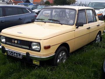 Een gele Wartburg 353 klassieke auto met een zwart kenteken met de tekst "85-04-UJ" valt op, geparkeerd op gras tussen moderne voertuigen.