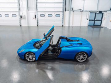 Een blauwe Gordon Murray Automotive T.33 Spider met de deur aan de bestuurderszijde open staat geparkeerd in een lege, schone inpandige garage met witte muren en grote gesloten garagedeuren.