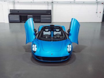 Een blauwe Gordon Murray Automotive T.33 Spider met open schaardeuren staat geparkeerd in een schone, moderne garage met zwarte kasten en laden langs de achterwand.