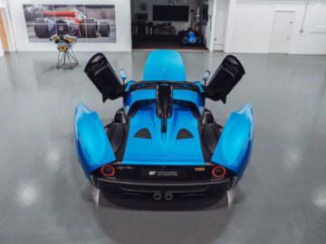 Een blauwe sportwagen met open vlinderdeuren in een moderne garage, met daarin een Gordon Murray Automotive T.33 Spider. Een foto van een raceauto siert de muur en er staat een tafel met bloemen vlakbij.