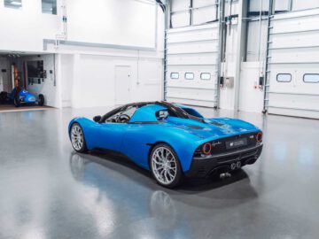 Een blauwe Gordon Murray Automotive T.33 Spider-sportwagen staat geparkeerd in een ruime, schone industriële garage met gesloten witte garagedeuren.