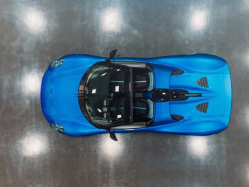 Bovenaanzicht van een strakke, blauwe Gordon Murray Automotive T.33 Spider met een glanzende afwerking, binnen geparkeerd op een reflecterende grijze vloer. De auto heeft een gestroomlijnd ontwerp en aerodynamische rondingen.