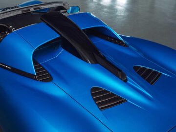 Close-up van de Gordon Murray Automotive T.33 Spider, een blauwe, hoogwaardige sportwagen met een gestroomlijnd ontwerp, zwarte aerodynamische vin en verschillende ventilatieopeningen aan de achterkant.