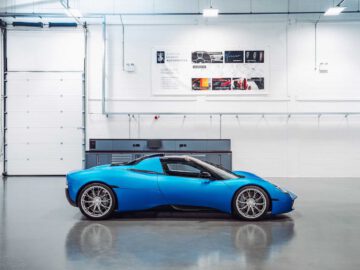 Een blauwe Gordon Murray Automotive T.33 Spider staat geparkeerd in een goed verlichte garage met witte muren, grote posters aan de muur en grijze kasten op de achtergrond.