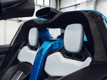 Binnenaanzicht van een blauwe Gordon Murray Automotive T.33 Spider converteerbare sportwagen met strakke, witleren stoelen met hoofdsteunen, een centrale bar en een modern dashboard in een goed verlichte garage.